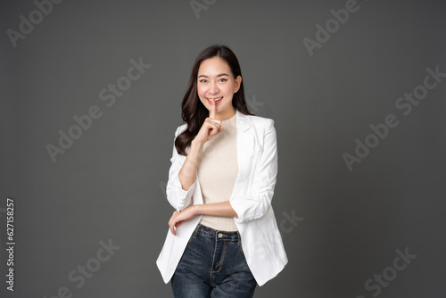 Asian female executive with long hair stand and pose Raise your hand and index finger to your mouth and smile. Wearing a white suit, jeans, making eye contact and standing in a gray screened studio.
