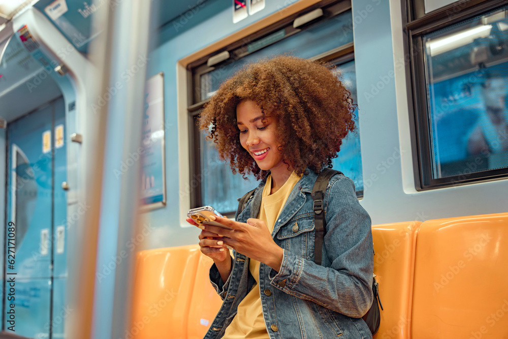 Naklejka premium Happy young African American woman passenger smile and using smart mobile phone in subway train station.