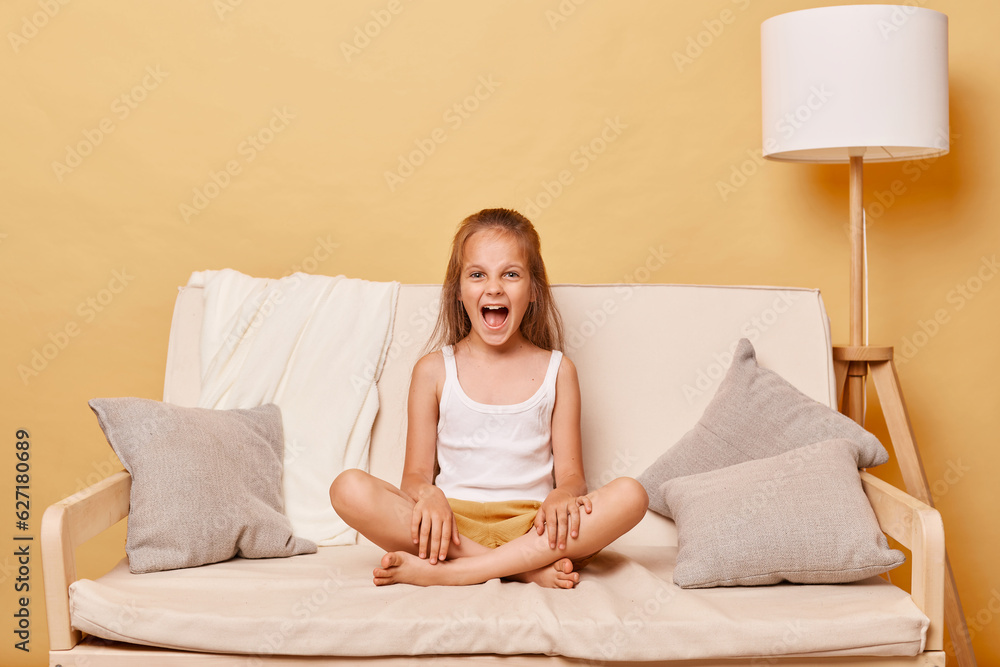 Φωτογραφία Angry little girl wearing casual clothing sitting on sofa at home against beige wall having problems during teenage years having hysterical behavior screaming