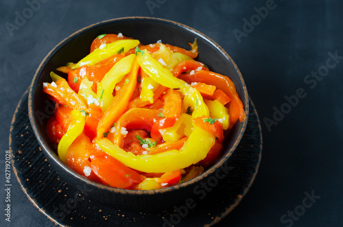 Salad with red and yellow roasted pepper slices, garlic and parsley in a black ceramic bowl on a black background. Concept of healthy eating. Traditional Italian dish. Vegetarian and vegan food.