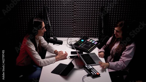 View from above of young woman podcaster interviewing her female guest, both wearing headphones and speaking into microphones in a modern acoustic studio 