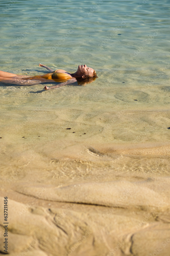 Obraz premium A young, sexy girl of European appearance, in a yellow bathing suit, lies in the sea, blue water on the beach, relaxes and sunbathes.