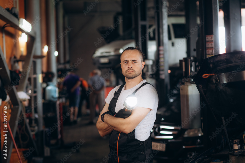 Handsome Car Mechanic is Posing in a Car Service. He Wears a Jeans Shirt and Safety Glasses. His Arms are Crossed. Specialist Looks at a Camera and Smiles Handsome Car Mechanic is Posing in a Car Service. He Wears a Jeans Shirt and Safety Glasses. His Arms are Crossed. Specialist Looks at a Camera and Smiles