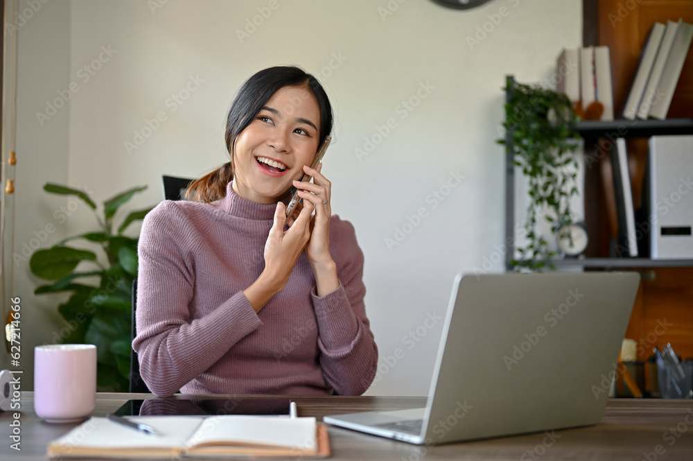 Cheerful young Asian woman having a phone call with her friend while working Cheerful young Asian woman having a phone call with her friend while working