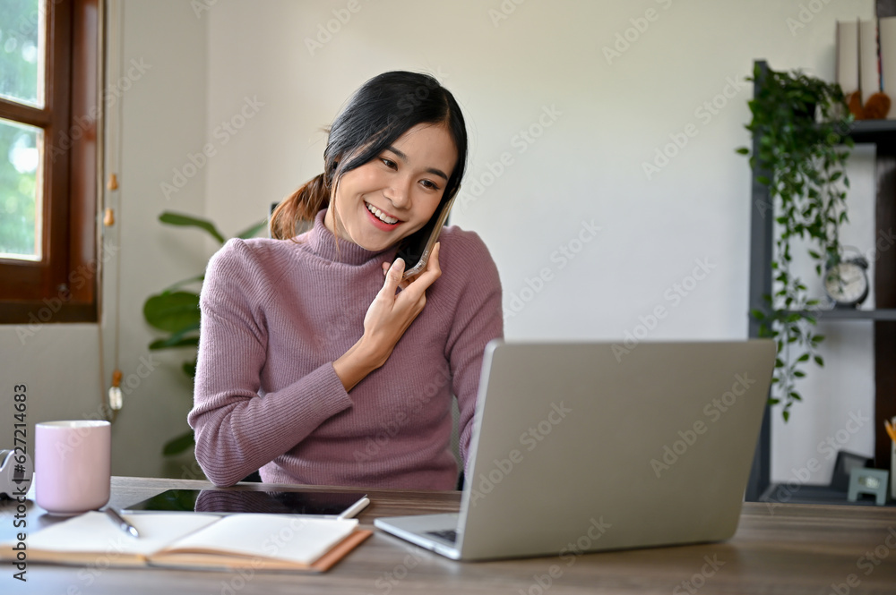 Cheerful young Asian woman having a phone call while working at her study table Cheerful young Asian woman having a phone call while working at her study table