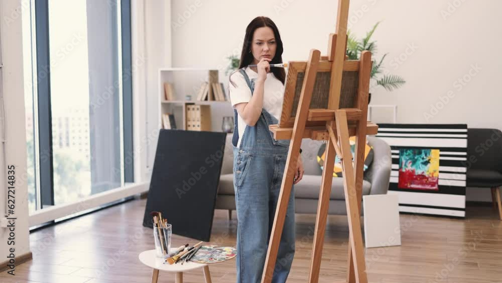 Attractive caucasian female applying brushstrokes on canvas supported by wooden easel indoors. Self-employed painter concentrating on play of light and shade while using comfort of art studio. Attractive caucasian female applying brushstrokes on canvas supported by wooden easel indoors. Self-employed painter concentrating on play of light and shade while using comfort of art studio.