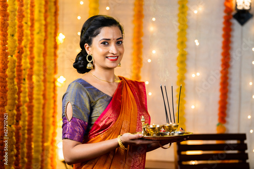 Happy indian girl in traditional ethnic wear holding pooja plate with incense sticks looking camera during diwali festival celebration at home - concept Indian culture, positive emotion and happiness photo