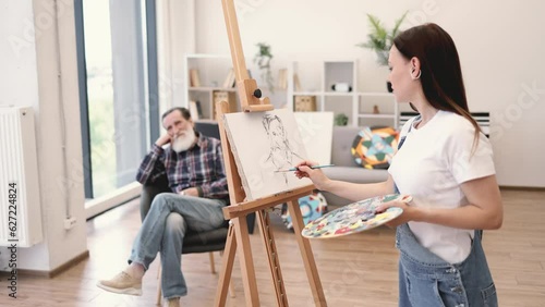 Self-employed female painter in denim overalls depicting sitter head-and-shoulders in modern studio space. Attractive brunette lady sketching out rough ideas of senior man's portrait on canvas. photo