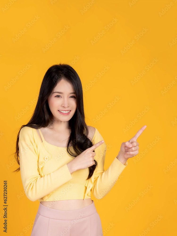 Beautiful Asian woman cute girl in with yellow shirt smiling and pointing finger to empty copy space for present product or blank space for advertising isolated on yellow background Beautiful Asian woman cute girl in with yellow shirt smiling and pointing finger to empty copy space for present product or blank space for advertising isolated on yellow background