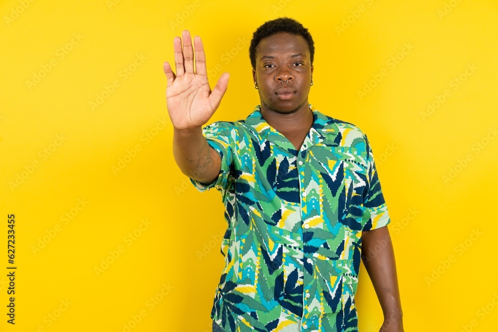 Young latin man wearing hawaiian shirt over yellow background doing stop sing with palm of the hand. Warning expression with negative and serious gesture on the face. Young latin man wearing hawaiian shirt over yellow background doing stop sing with palm of the hand. Warning expression with negative and serious gesture on the face.