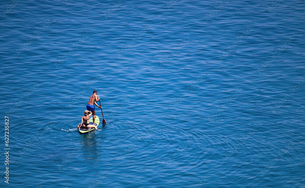A young white Caucasian couple paddle boarding on the calm blue sea - man standing pushing an oar into the water, woman sitting on the board - high-angle overhead drone shot with copyspace for text