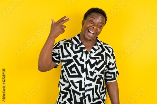  Young latin man wearing printed shirt over yellow background foolishness around shoots in temple with fingers makes suicide gesture. Funny model makes finger gun pistol