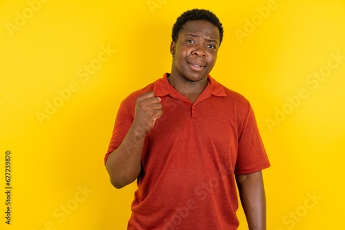 Young latin man wearing red t-shirt over yellow background shows fist has annoyed face expression going to revenge or threaten someone makes serious look. I will show you who is boss photo