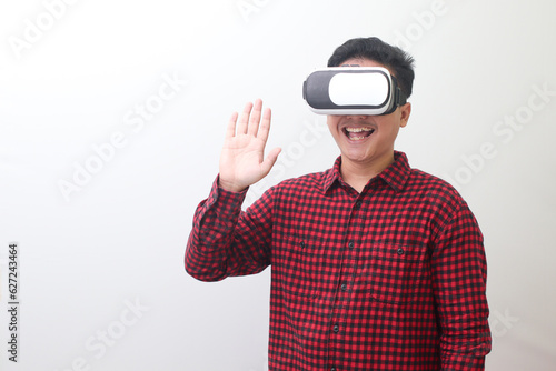 Portrait of Asian man in red plaid shirt using Virtual Reality (VR) glasses and greet his friend inside the game with opened palm. Isolated image on white background