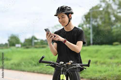 Fototapeta Naklejka Na Ścianę i Meble -  Smiling cyclist in sport clothes sitting on his bike and using mobile phone. Sport, technology and active lifestyle concept