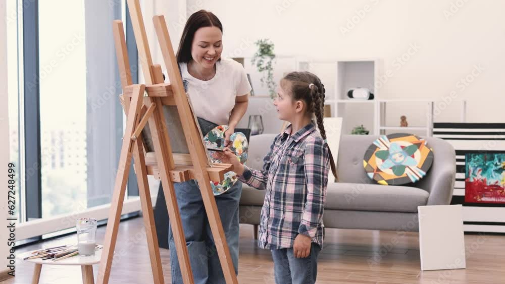 Portrait of beautiful female and little child in casual clothes standing behind easel with painting in workshop. Attentive art person teaching inquisitive small student mixing oil paints on paper. Portrait of beautiful female and little child in casual clothes standing behind easel with painting in workshop. Attentive art person teaching inquisitive small student mixing oil paints on paper.