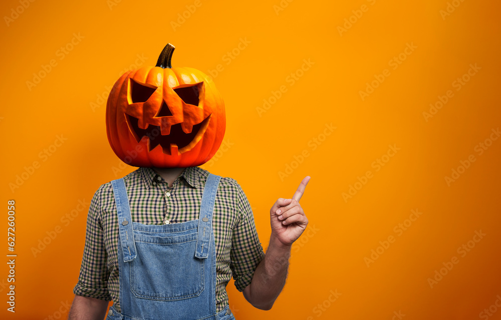 Person in Halloween costume of scarecrow with Jack-o'-lantern pumpkin head points away over orange background with copy space - obrazy, fototapety, plakaty 
