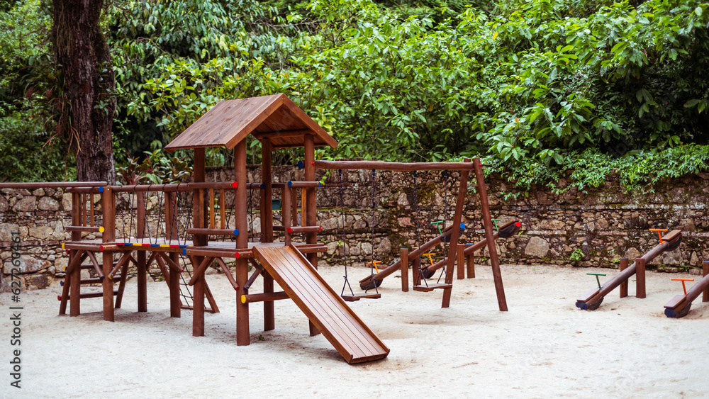 View of a children's playground in a tropical park with a group of seesaws in the background next to the stone wall, wooden kid's slide and swing in front of the teeter-totters on the sand View of a children's playground in a tropical park with a group of seesaws in the background next to the stone wall, wooden kid's slide and swing in front of the teeter-totters on the sand