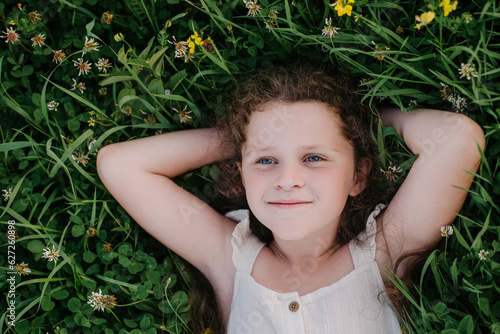 Top view of relaxed smiling adorable little girl kid lying on fresh green grass in park at sunny day. Carefree pretty child lie on field and enjoying summer weekend. Nature and childhood concept