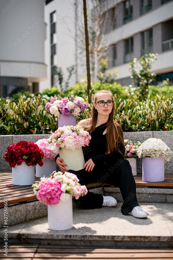 Fototapeta premium Cute young girl surrounded with amazing bouquets with fresh flowers arrangement in hat boxes on blurred street background