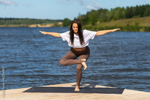 Woman practicing yoga, performs the exercise Eka Pada Agnistambha Utkatasana, balance asana, trains in sportswear while standing on one leg on a wooden bridge on the shore of the lake