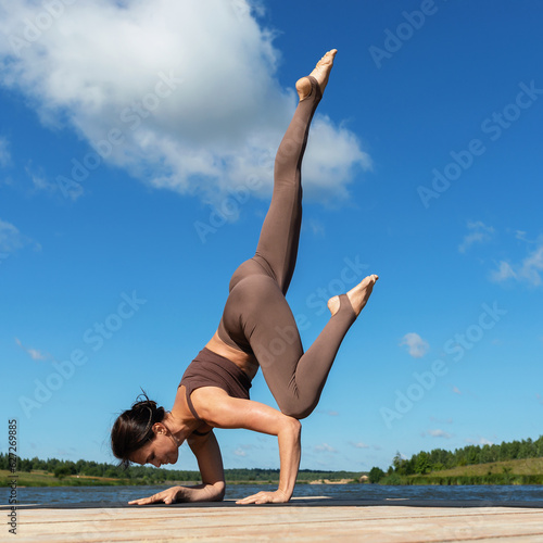 A woman practicing yoga, performs the exercise Eka Pada Bakasana, the pose of a crane with a leg extended up, trains in sportswear while standing on her hands on a wooden bridge on the shore of a lake