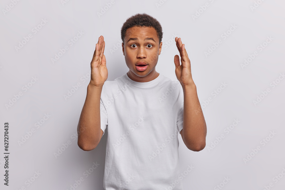 Φωτογραφία Astonished surprised dark skinned man with short curly hair wears casual t shirt demonstrates big size huge object holds breath from amazement poses against white background