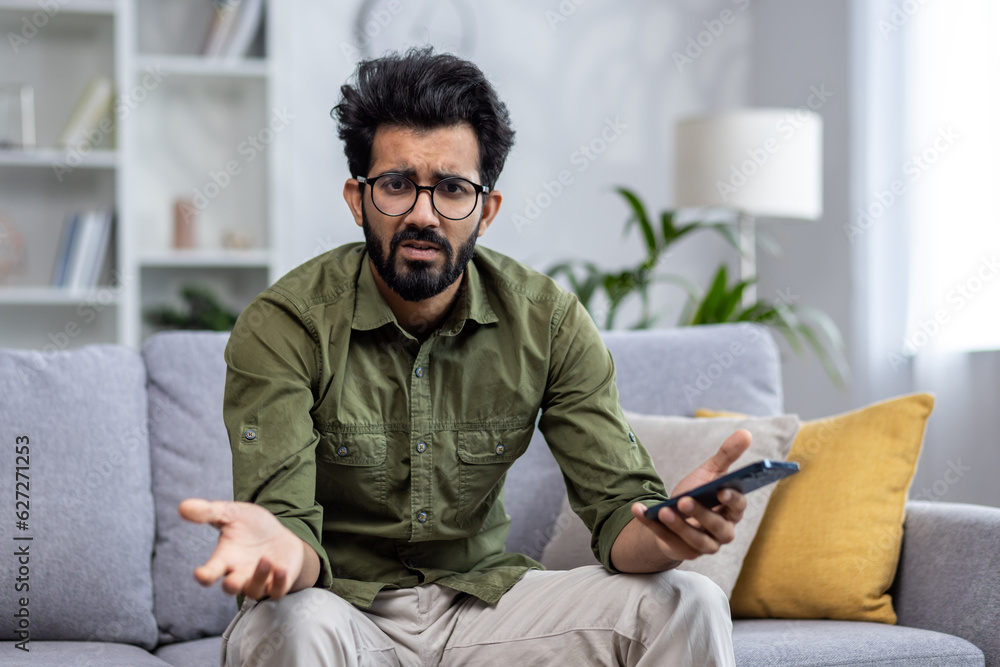 Portrait of unhappy man at home, indian man upset and disappointed sitting on sofa in living room and looking at camera, holding phone.