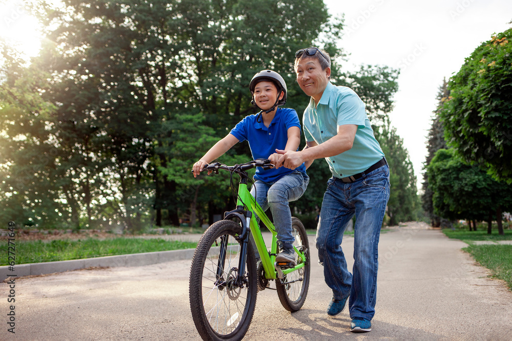 asian dad teaches son to ride bike, korean senior helps child, boy in helmet is actively relaxing with his father asian dad teaches son to ride bike, korean senior helps child, boy in helmet is actively relaxing with his father