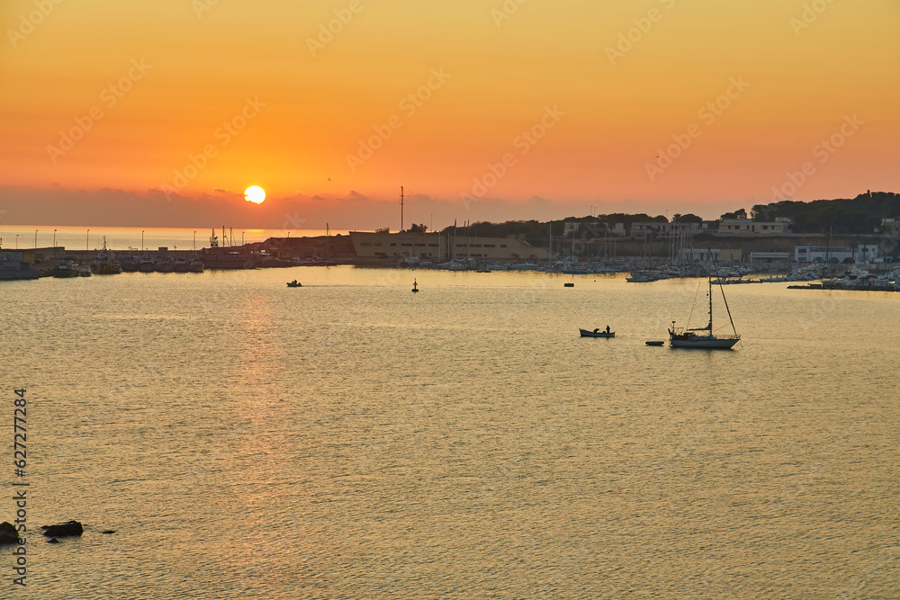 beautiful orange sunset or sunrise over the harbor of Otranto, Italy. Soft clouds, calm ocean and backlit skyline and marina with yachts and fishermen.