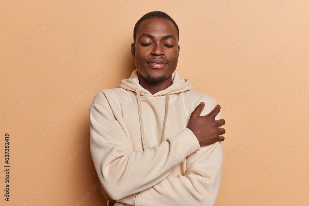 Serene dark skinned man dressed in casual brown sweatshirt embraces himself has eyes closed finding comfort and solace epitomizes inner peace and self acceptance isolated over beige background Serene dark skinned man dressed in casual brown sweatshirt embraces himself has eyes closed finding comfort and solace epitomizes inner peace and self acceptance isolated over beige background