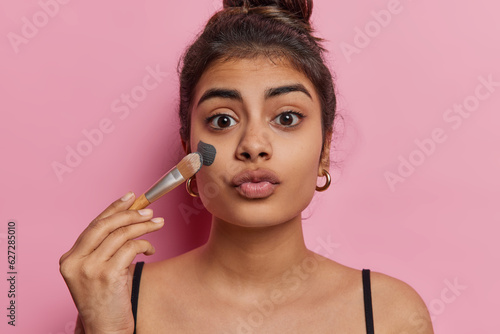 European woman applies beauty mask with cosmetic brush embracing her skincare routine with delight embodying bliss of beauty procedures keeps lips rounded dressed in t shirt isolated on pink wall