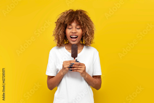 young curly american girl with braces eats chocolate ice cream on yellow isolated background