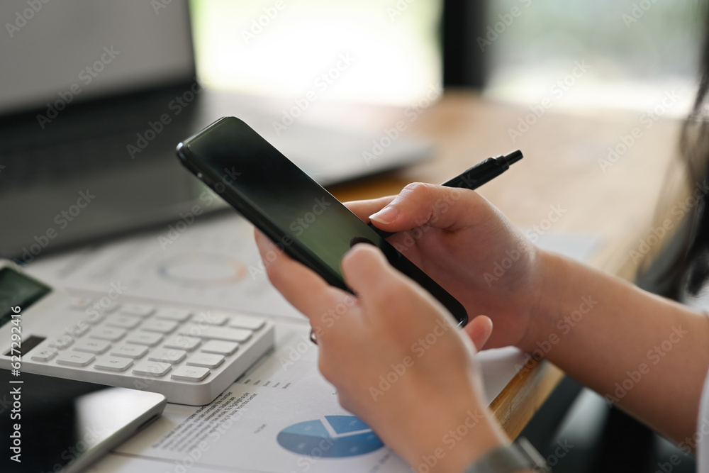 Fototapeta premium Close up with female hand holing smart phone and touching on device screen while doing a financial documents on digital tablet, business data analysis concept.