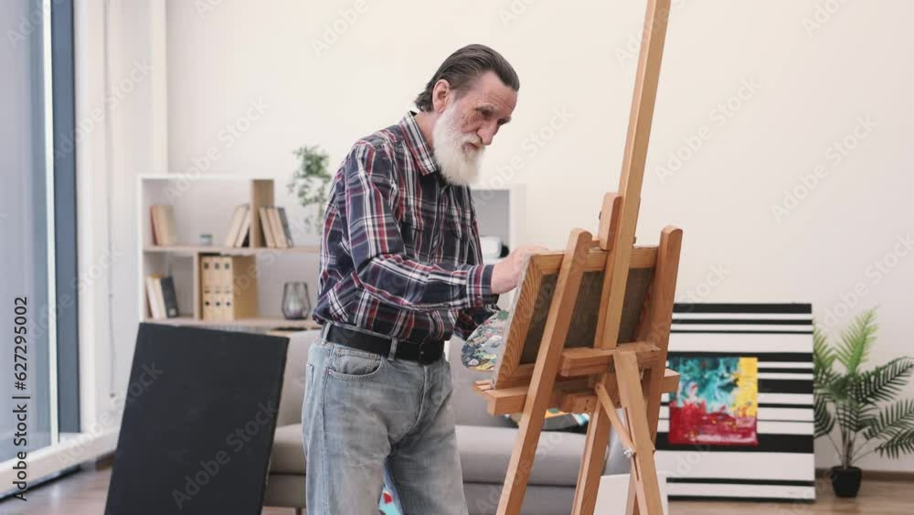 Close up view of elderly male in denim wear with palette layering paint on cloth with brush in studio interior. Involved person honing professional skills and work in designated area of art space. Close up view of elderly male in denim wear with palette layering paint on cloth with brush in studio interior. Involved person honing professional skills and work in designated area of art space.