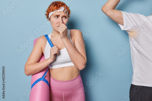 Unhappy redhead sportswoman instinctively covers her nose in attempt to escape stench avoids unbearable smell dressed in sportswear with karemat looks at sweaty guy with raised arm. Sport concept