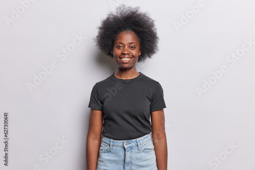 Charming display of positive emotions. Smiling dark skinned woman with bushy hair emanates joy as she smiles graciously dressed in casual black tshirt and jeans poses in studio over white background