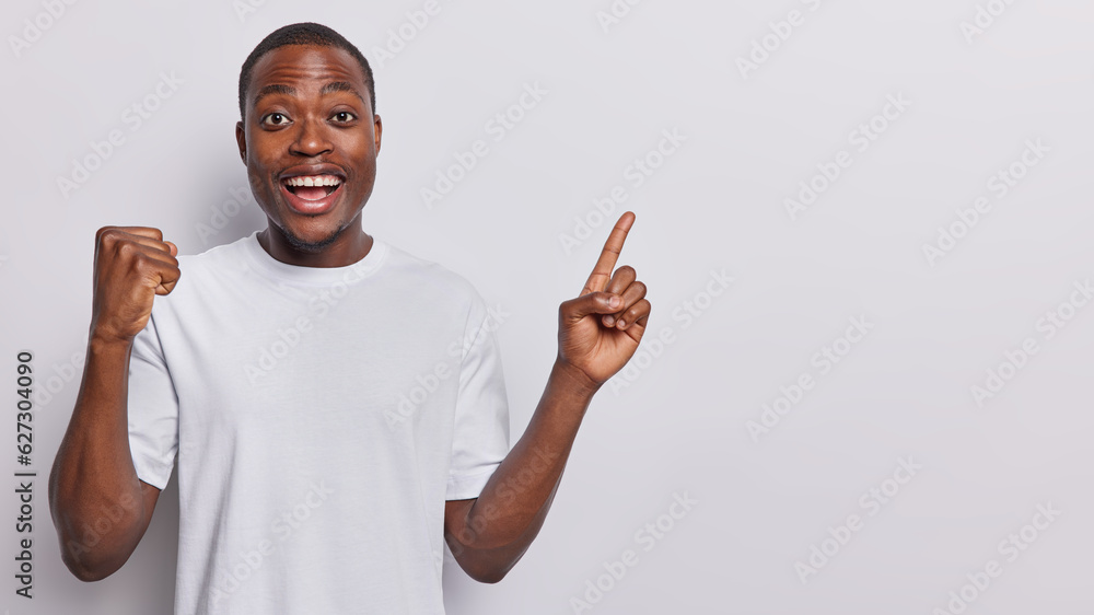 Waist up shot of positive dark skinned man clenches fist with triumph celebrates good news and nice offer points index finger at empty space for your promotional text isolated over white background Waist up shot of positive dark skinned man clenches fist with triumph celebrates good news and nice offer points index finger at empty space for your promotional text isolated over white background