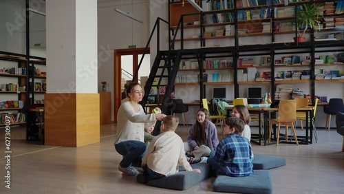 Smiling teacher tapes papers to foreheads of children for game of who am I sitting in circle on pillows on floor in library 