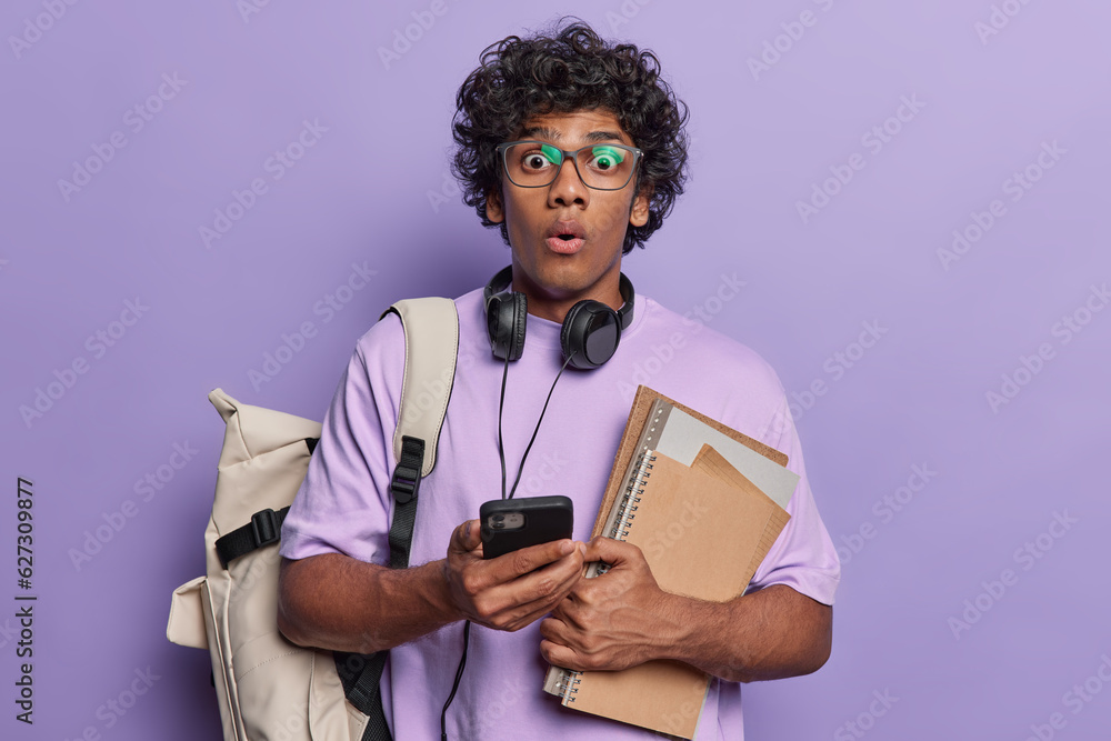 Impressed curly haired Hindu student holding books and notepads carrying backpack uses mobile phone for chatting with friends dressed in tshirt isolated on purple. Education school college concept Impressed curly haired Hindu student holding books and notepads carrying backpack uses mobile phone for chatting with friends dressed in tshirt isolated on purple. Education school college concept
