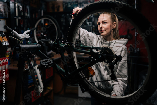 Attractive skilled cycling repairman female repairing and fixing mountain bicycle standing on bike rack in repair workshop with dark interior. Concept of professional maintenance of bicycle transport.