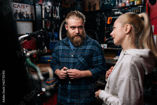 Professional fashion cycling repairman with beard communicating with pretty blonde female client, talking problem of bicycle, detected during diagnostics in repair shop with dark interior.