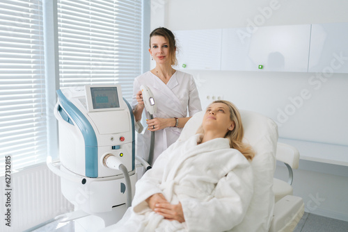 Portrait of female beautician specialist standing posing with device for photo rejuvenation in cosmetology clinic  looking at camera. Woman client receiving stimulating electric facial treatment.