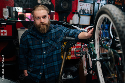 Portrait of serious bearded cycling mechanic male standing by bicycle in repair bike workshop with dark interior, looking at camera. Concept of professional repair and maintenance of bicycle transport