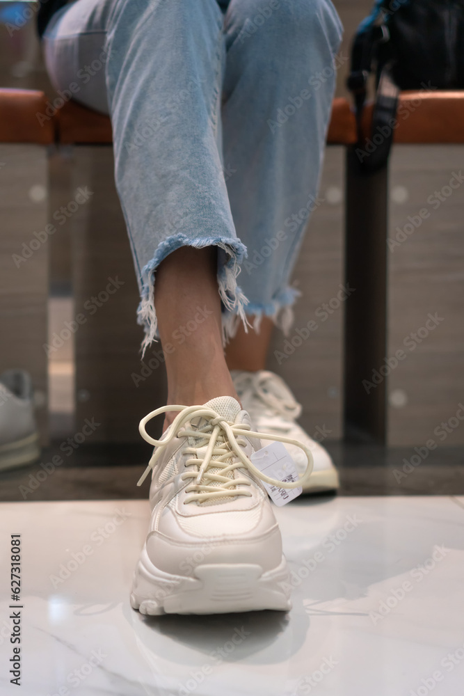 Close-up of women's legs choosing sports shoes and trying on different white sneakers at the mall. Women's hands close-up tie shoelaces when trying on sports shoes. Vertical photo