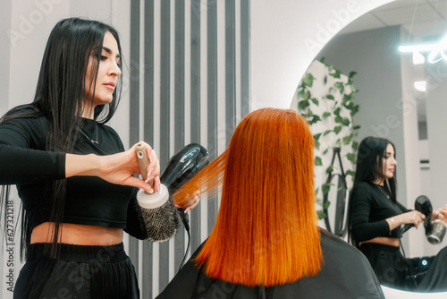 Woman hairdresser does combing red hair to client and does hairstyle in beauty salon in front of mirror. Hair styling and straightening.