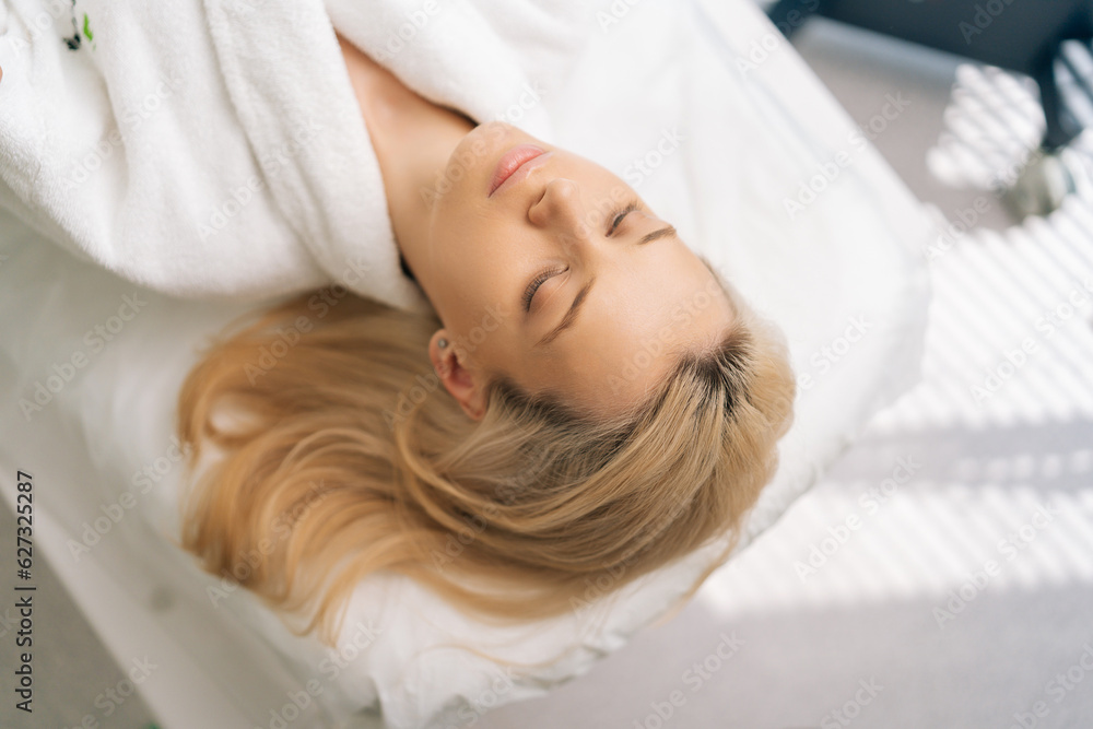 Close-up top view of serene blonde female wearing white bathrobe lying with closed eyes on medical couch after spa treatment. Close up face of happy young woman relaxing enjoying daily hygiene routine Close-up top view of serene blonde female wearing white bathrobe lying with closed eyes on medical couch after spa treatment. Close up face of happy young woman relaxing enjoying daily hygiene routine