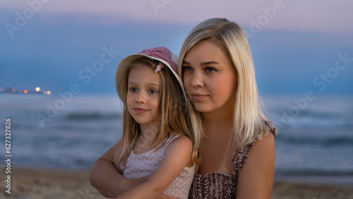 Mother and daughter on beach of sea or ocean in evening at sunset. Concept of summer vacation, travel, holiday. People outdoors on seascape
