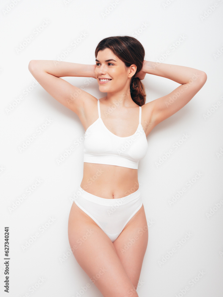 Obraz premium Portrait of young beautiful sexy woman in white lingerie. Smiling carefree model wearing pure underwear. Hot brunette posing near wall in studio interior. Perfect body. Slim and fit
