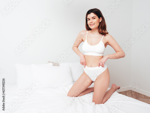 Attractive and relaxed seductive young woman with slim body wearing white underwear. Sexy smiling model lying on sheets in the bedroom at home in the early morning in lingerie. Happy female in bed